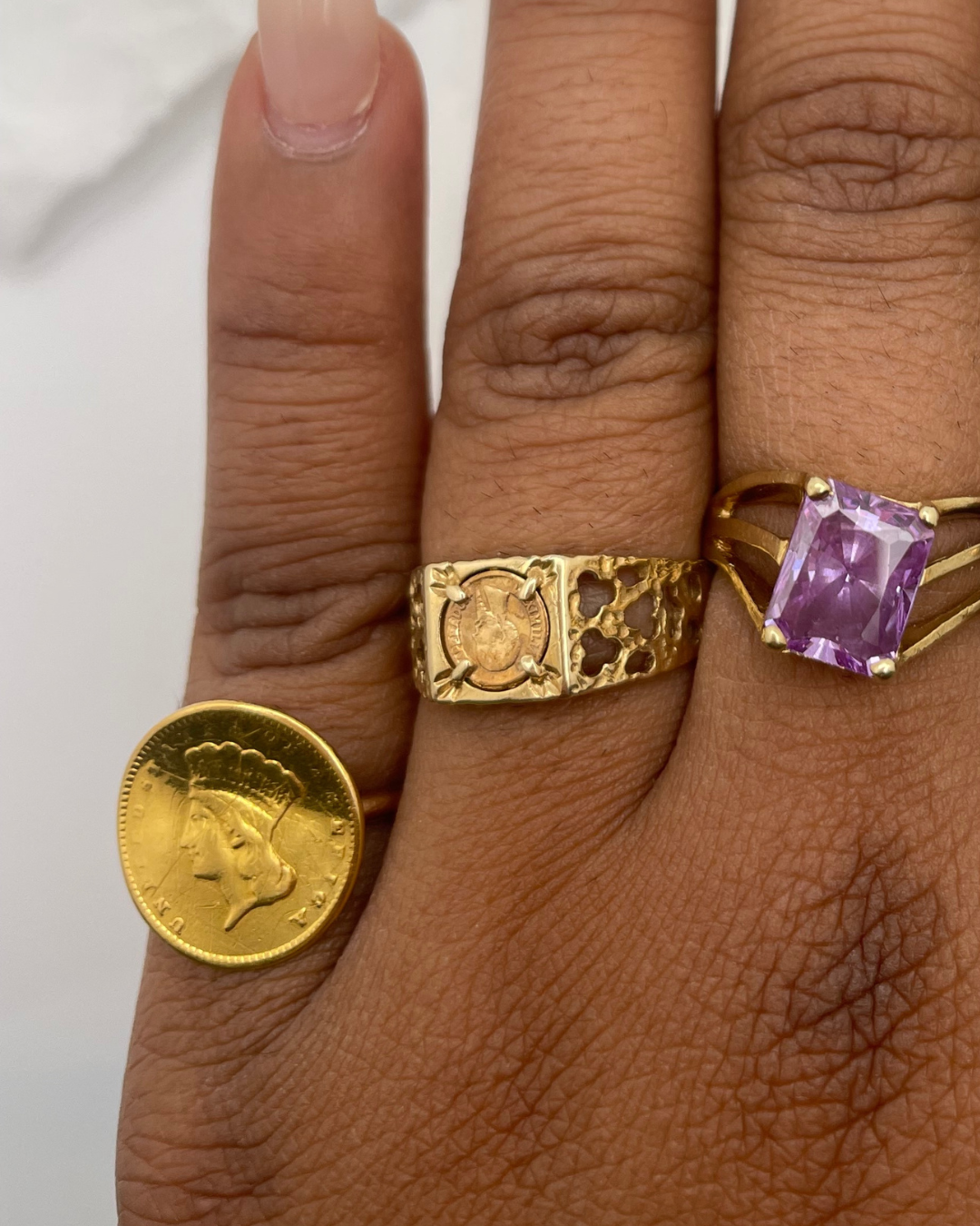 Close-up of a hand wearing two gold rings with a coin and purple gemstone on a white background